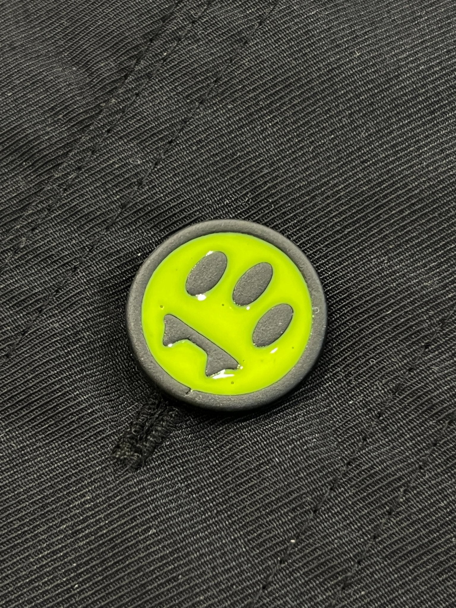 A round pin with a neon yellow-green background and black sad face is attached to black fabric, giving the BARROW S5BWUAJK065 Jacket Unisex Nero/Black an edgy urban accent perfect for accessorizing your look.