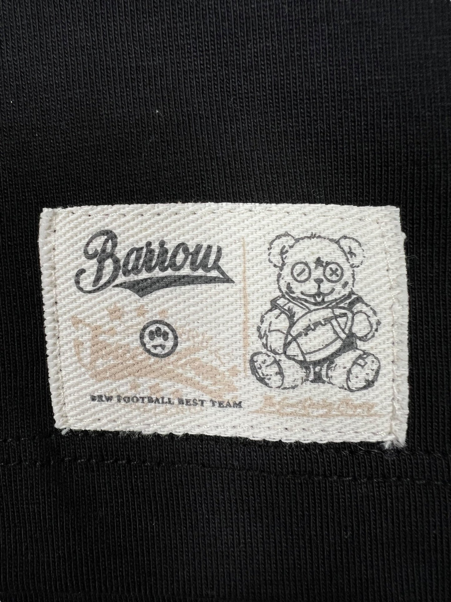 The cloth label on the black Barrow S5BWUATH022 Jersey T-shirt by BARROW features "Barrow" text, a bear with a football, and the phrase "New Football Best Team," made from 100% cotton.