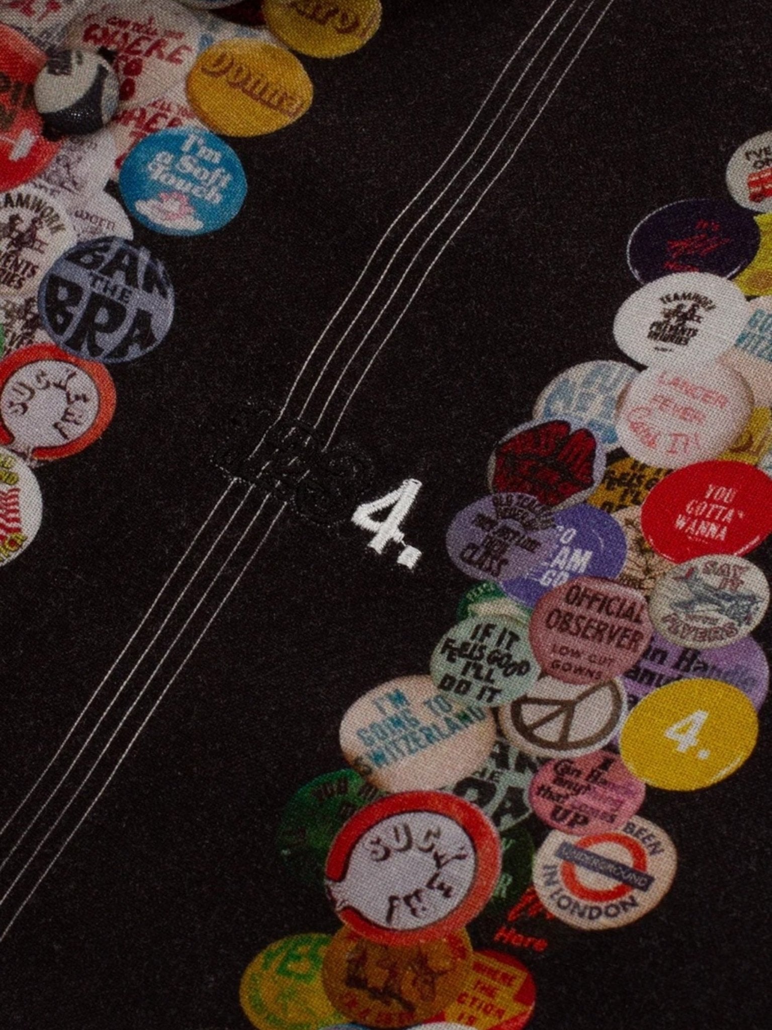 A close-up of the BLDG4 I love Bldg4 SS Woven Black shirt by BLDG4 shows colorful round pins with various designs and stitched white lines around the number "4," adding unique flair to this linen rayon piece.