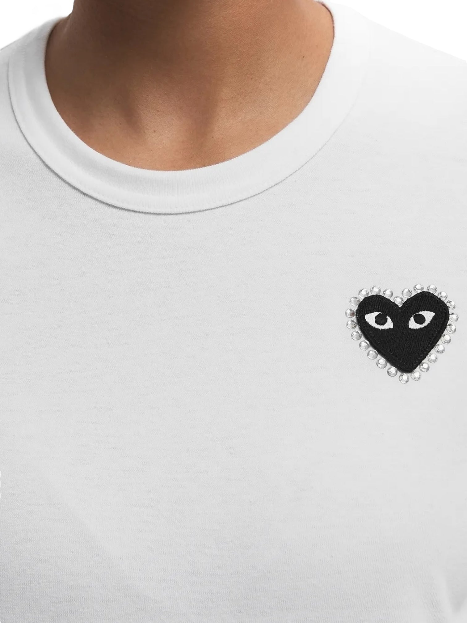 Person wearing a Comme des Garcons P1T801 T-SHIRT RHINESTONE HEART WHT/BLK by COMME DES GARCONS X PLAY, featuring a black heart patch with eyes and rhinestone border on the left chest.