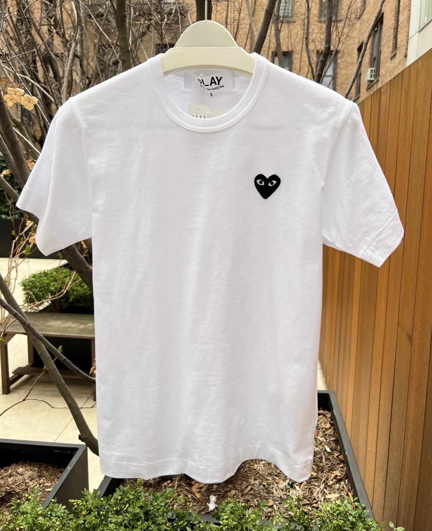 COMME DES GARÇONS P1T064 PLAY T-Shirt with a small, black heart emblem hanging on a hanger outdoors, near a wooden fence and potted plants. The embroidered logo adds a touch of Comme des Garçons style to your wardrobe.