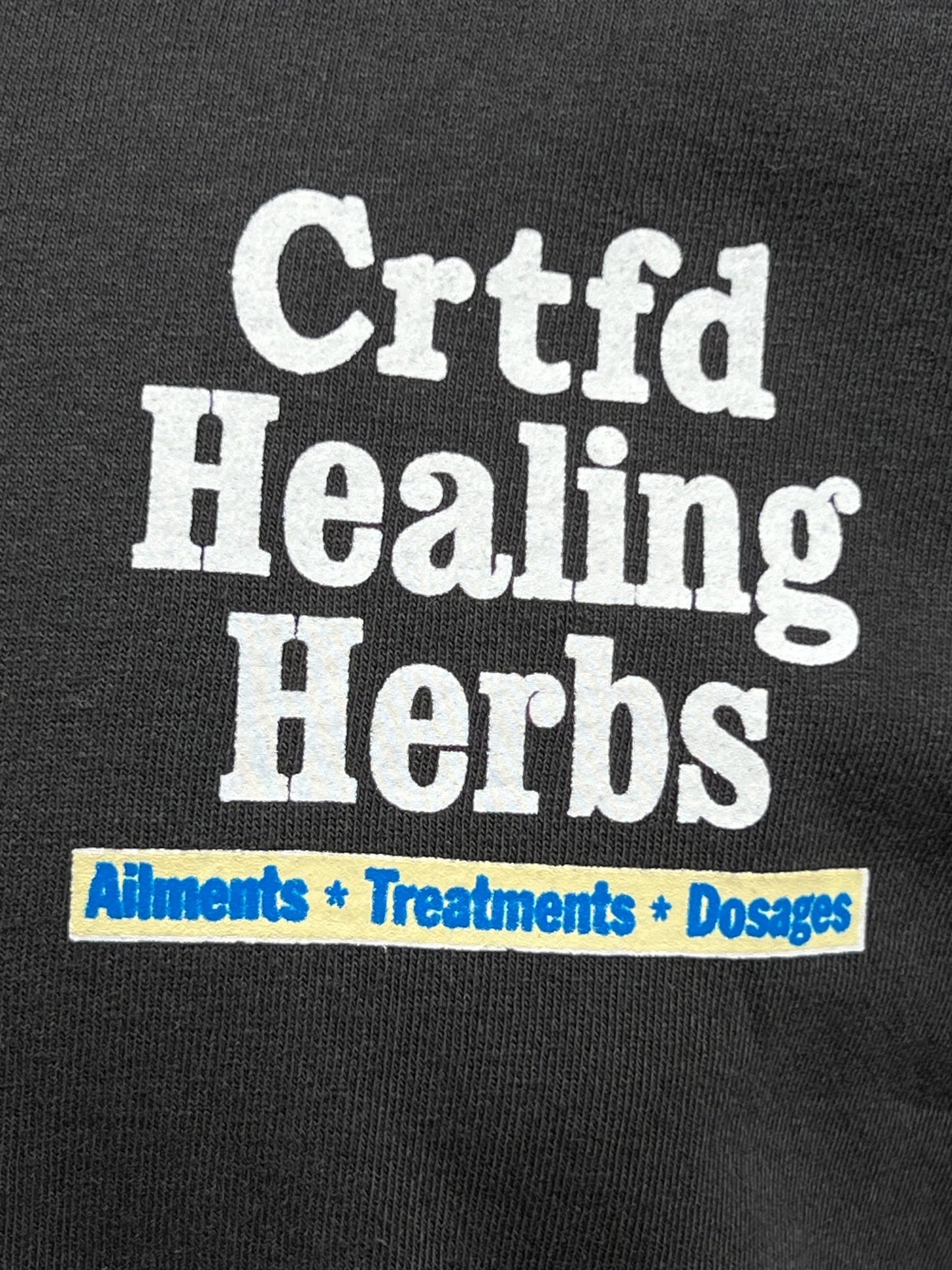 Close-up of a text design on a black, organic cotton CRTFD AYURVEDIC T-SHIRT by CRTFD that reads, "Crtfd Healing Herbs," followed by "Ailments * Treatments * Dosages" in smaller text below.