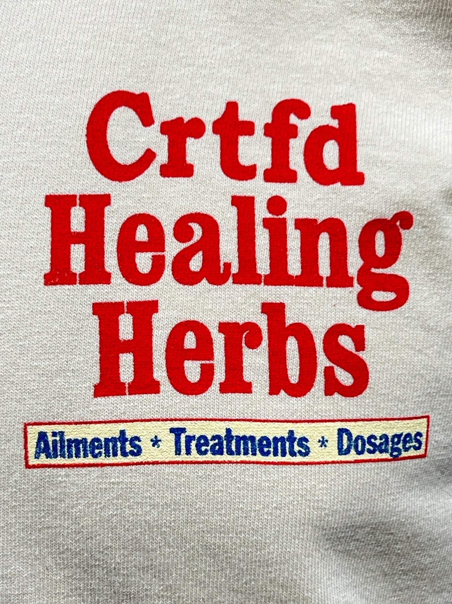 Close-up of a logo with red text reading "Crtfd Healing Herbs" on an organic cotton, beige CRTFD Ayurvedic T-Shirt Natural, and a blue section below listing "Ailments * Treatments * Dosages.