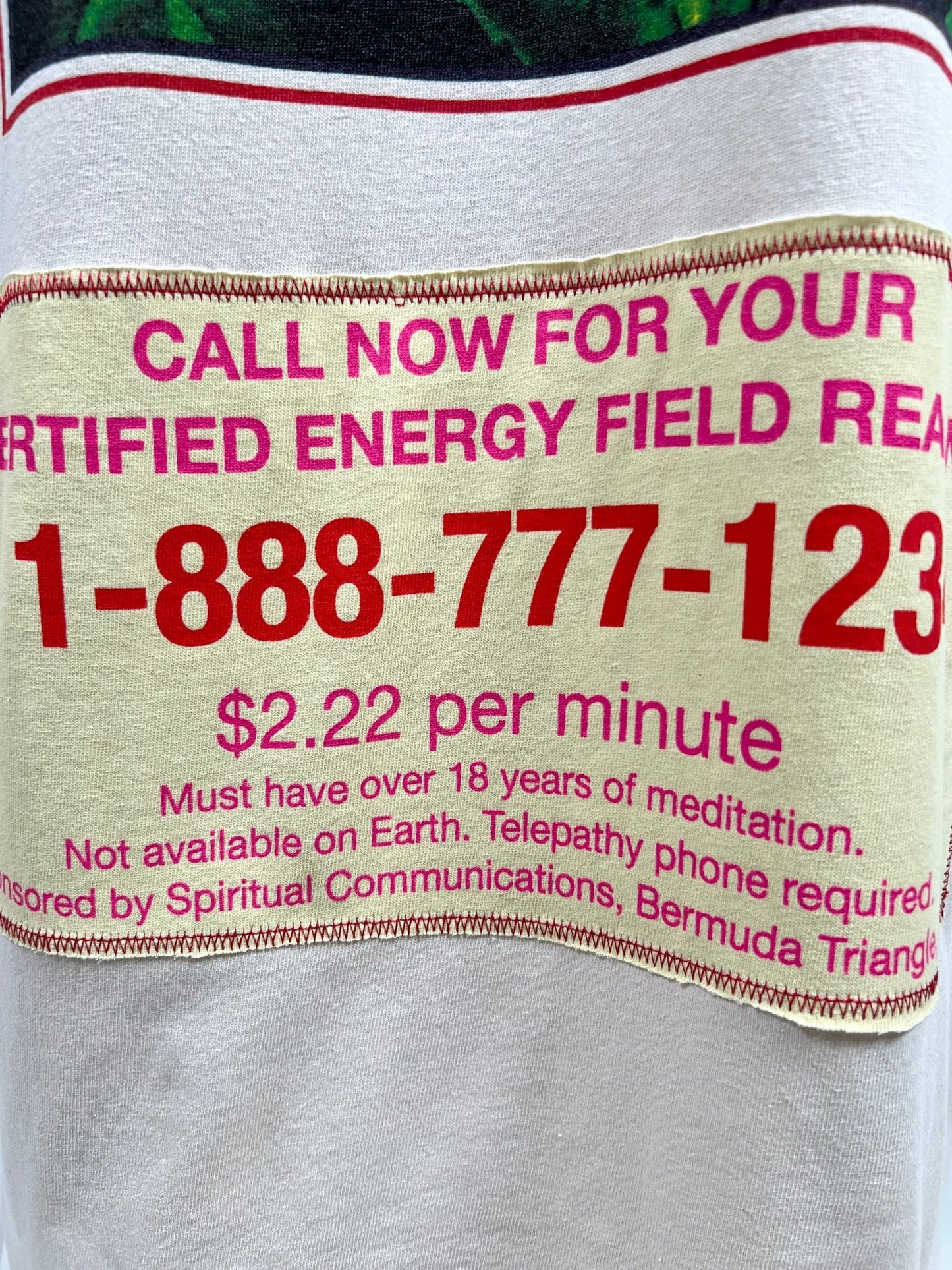 Close-up of a beige sign advertising a certified energy field reading for $2.22 per minute. Requires over 18 years of meditation and telepathy phone. Sponsored by Spiritual Communications, Bermuda Triangle. The reader wears a CRTFD AYURVEDIC T-SHIRT NATURAL made from 100% organic cotton for authenticity and comfort.