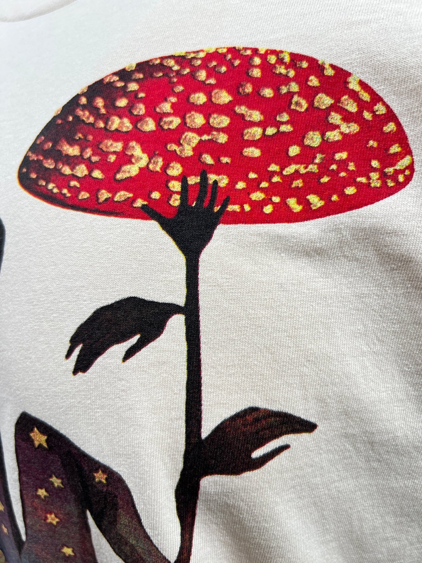 Close-up of a natural CRTFD FLY AGARIC T-SHIRT from CRTFD, featuring an abstract design with a red and yellow speckled mushroom and silhouetted leaves, crafted from 100% organic cotton.