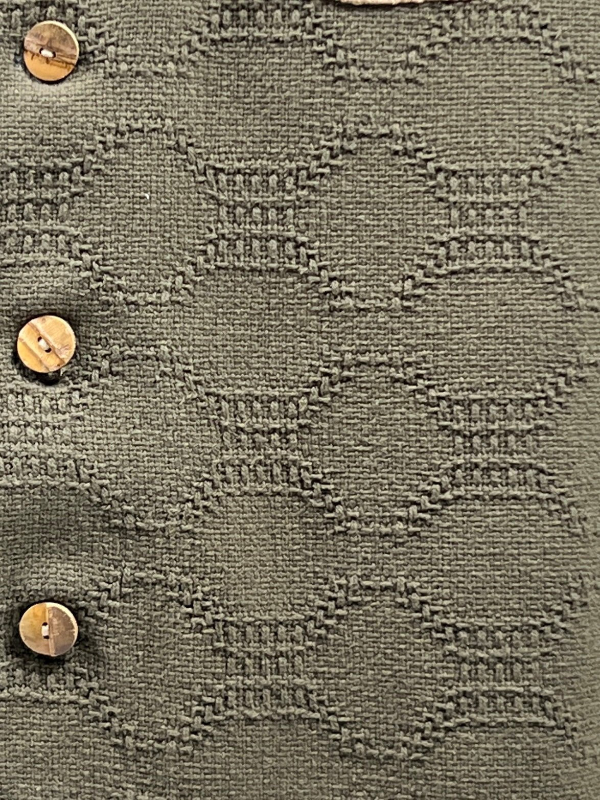 Close-up of a textured fabric with a repeating abstract pattern in dark, earthy olive green. Four wooden buttons adorn the 100% Organic Cotton CRTFD M51 Combo Shirt Olive by CRTFD.