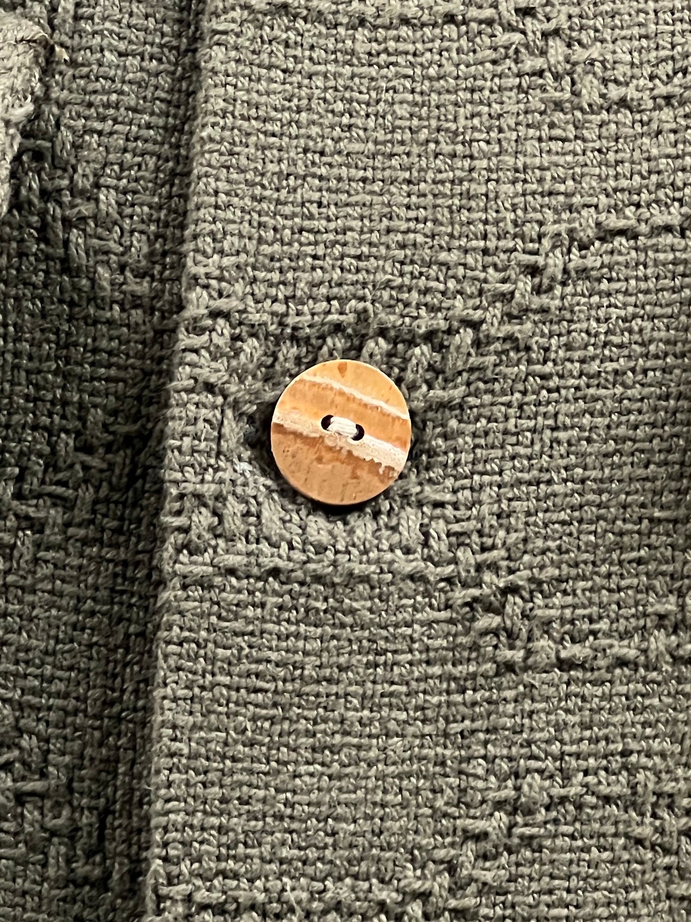 A close-up of a tan button sewn onto an olive green, textured fabric. The material appears to be woven with a rough texture and the buttonhole is visible, suggesting it's part of the CRTFD M51 COMBO SHIRT OLIVE by CRTFD.