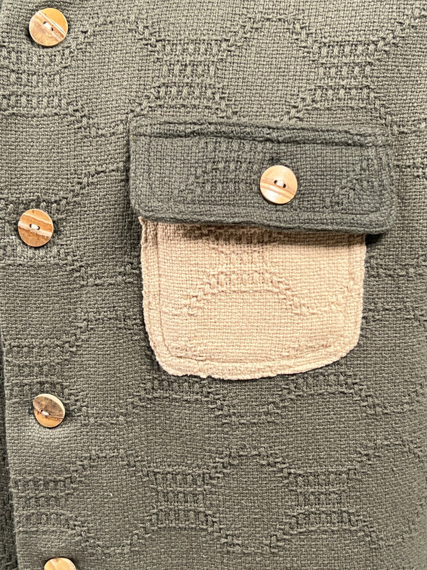 Close-up of the CRTFD M51 COMBO SHIRT OLIVE by CRTFD, featuring a textured greenish fabric with a beige pocket and wooden buttons, showcasing a woven pattern on the 100% organic cotton material.