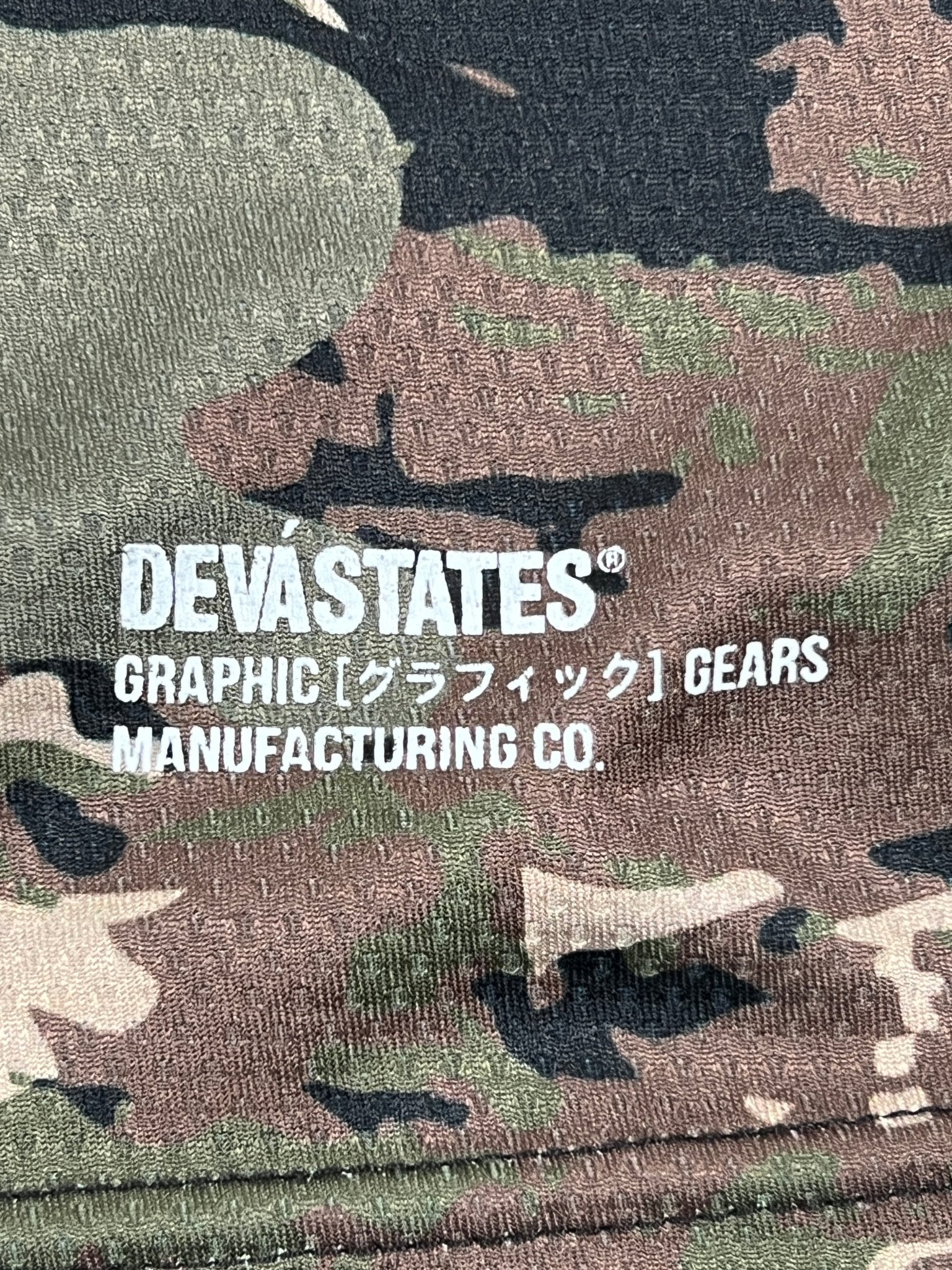 The Deva'States Ballistic s/s Mesh Jersey Top Camo by DEVÁ STATES is a polyester camo mesh jersey with white "DEVA STATES® GRAPHIC [グラフィック] GEARS MANUFACTURING CO." print.
