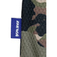 Close-up of the Deva'States Ballistic s/s Mesh Jersey Top Camo by DEVÁ STATES, featuring polyester camouflage fabric and a small purple tag with "DENÁ" in white, echoing the brand’s signature design.