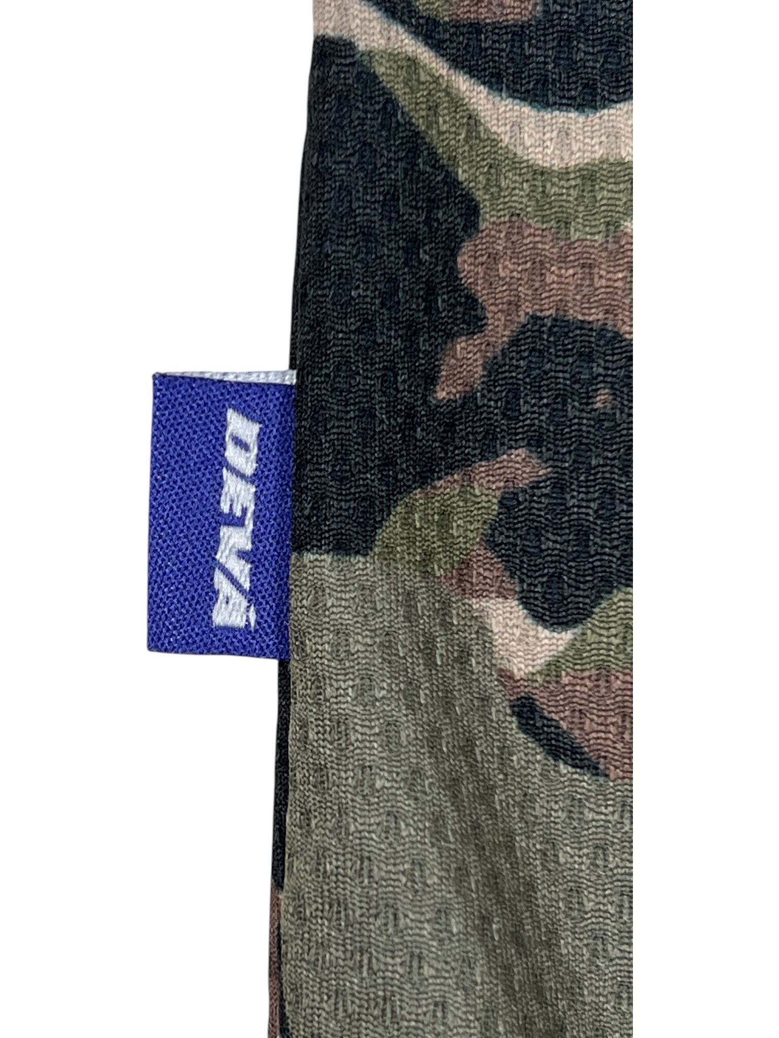 Close-up of the Deva'States Ballistic s/s Mesh Jersey Top Camo by DEVÁ STATES, featuring polyester camouflage fabric and a small purple tag with "DENÁ" in white, echoing the brand’s signature design.