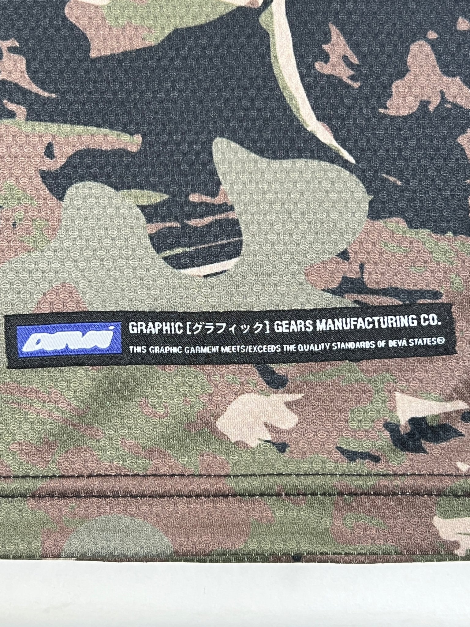 Close-up of camouflage-patterned polyester fabric with a "GRAPHIC GEARS MANUFACTURING CO." label. Perfect to pair with the DEVÁ STATES Deva'States Ballistic s/s Mesh Jersey Top Camo.