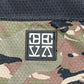 A black patch with white abstract symbols is sewn onto camouflage polyester fabric below a solid black section, giving the DEVÁ STATES Ballistic s/s Mesh Jersey Top Camo its signature edge.