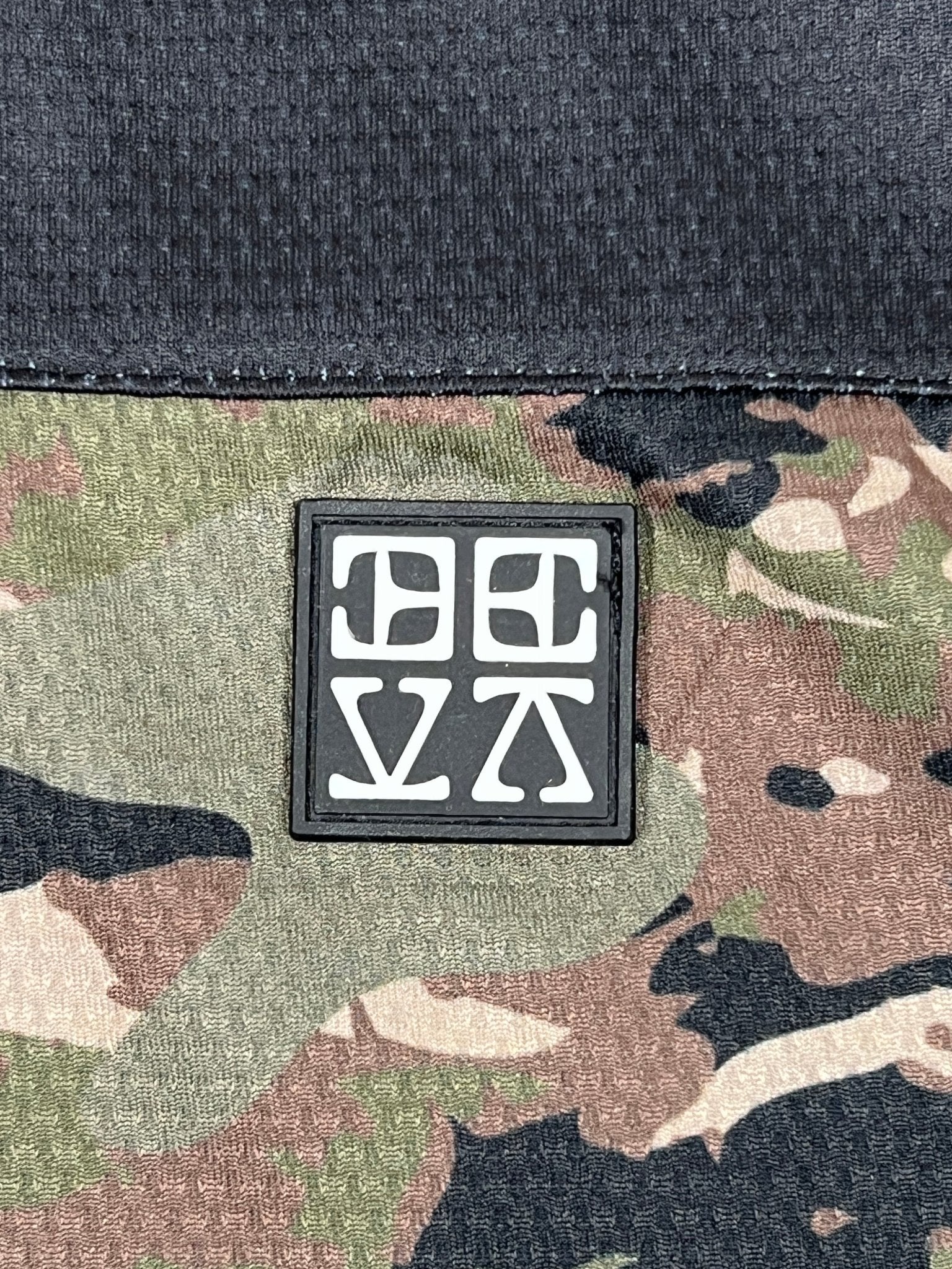 A black patch with white abstract symbols is sewn onto camouflage polyester fabric below a solid black section, giving the DEVÁ STATES Ballistic s/s Mesh Jersey Top Camo its signature edge.