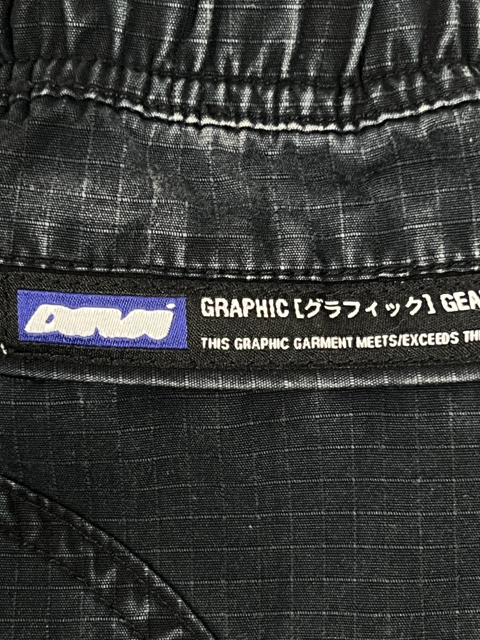 Close-up of a black fabric tag reading "GRAPHIC [グラフィック] GEAR" and "THIS GRAPHIC GARMENT MEETS/EXCEEDS TH," attached to DEVA'STATES Deva'States Hike Cargo Shorts in washed black cotton with a vintage washed finish.