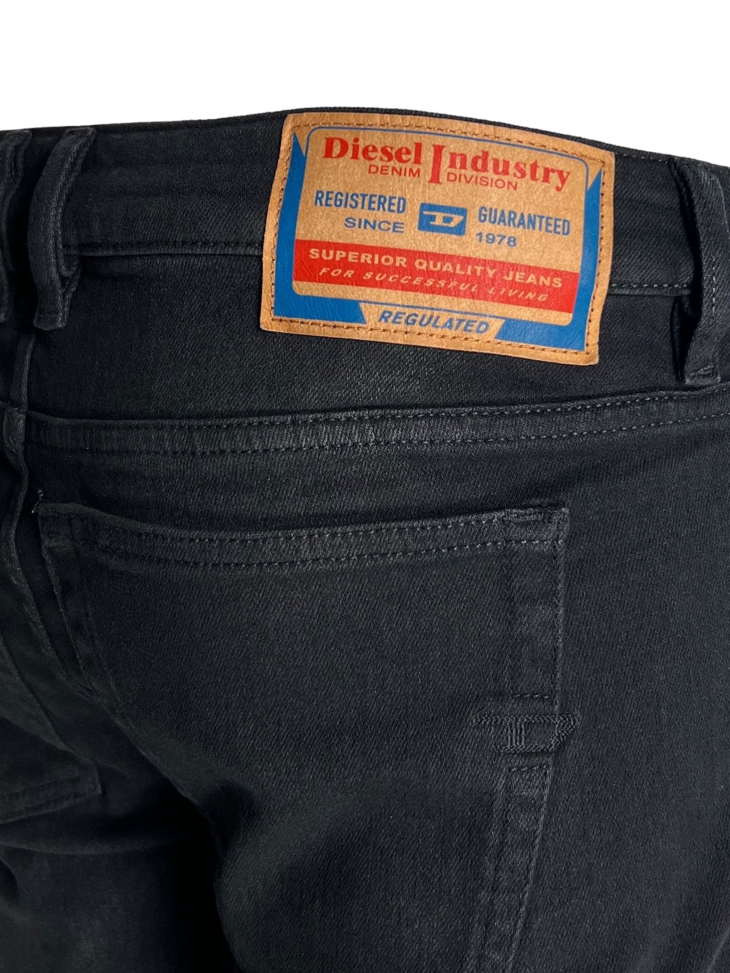 Close-up image of the back of black skinny style DIESEL 1979 SLEENKER 9H94 DENIM BLACK showing a brown label with "Diesel Industry" branding, indicating registration since 1978, and stating "Superior Quality Jeans" with blue and red details. The stretch denim fabric adds comfort while maintaining a punk-rock attitude.