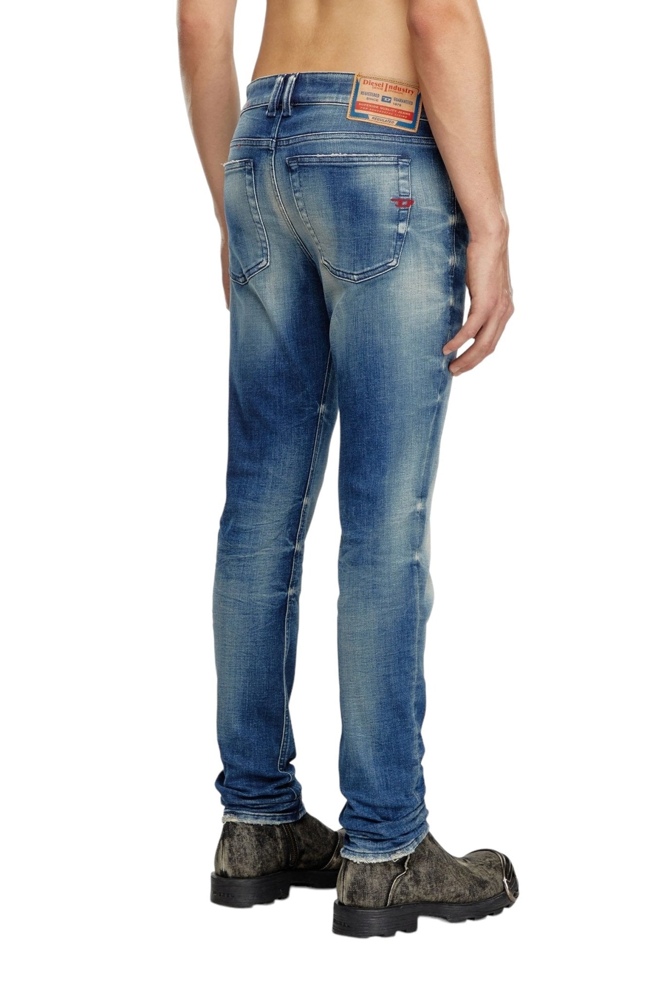 A person is shown from the back wearing DIESEL 1979 SLEENKER 9J24 medium blue stretch denim jeans and brown boots, exuding a punk-rock attitude. The jeans, made from organic cotton by the brand DIESEL, have a label on the waistband.