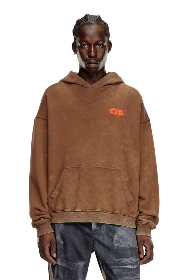 A person with braided hair is wearing the DIESEL S-BOXT-HOOD-Q5 brown Men's hooded sweatshirt from DIESEL, crafted from organic cotton with a small orange graphic on the upper left chest, paired with distressed dark pants.