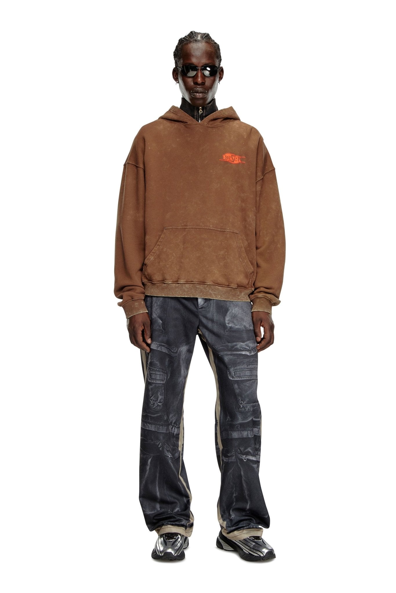 A person wearing the DIESEL S-BOXT-HOOD-Q5 BROWN, a relaxed fit men's hooded sweatshirt made from organic cotton, dark blue patterned pants, black shoes, and dark sunglasses stands against a plain white background.