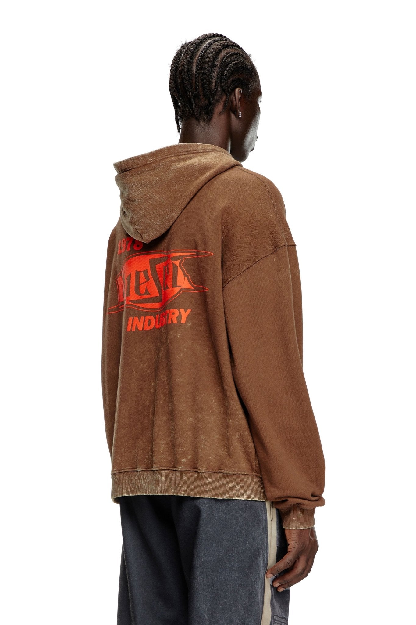 A person with braided hair is wearing the DIESEL S-BOXT-HOOD-Q5 BROWN, a relaxed fit hoodie made from organic cotton by DIESEL. The hoodie, which features red text on the back, is paired with loose-fitting pants. The individual is facing away from the camera.