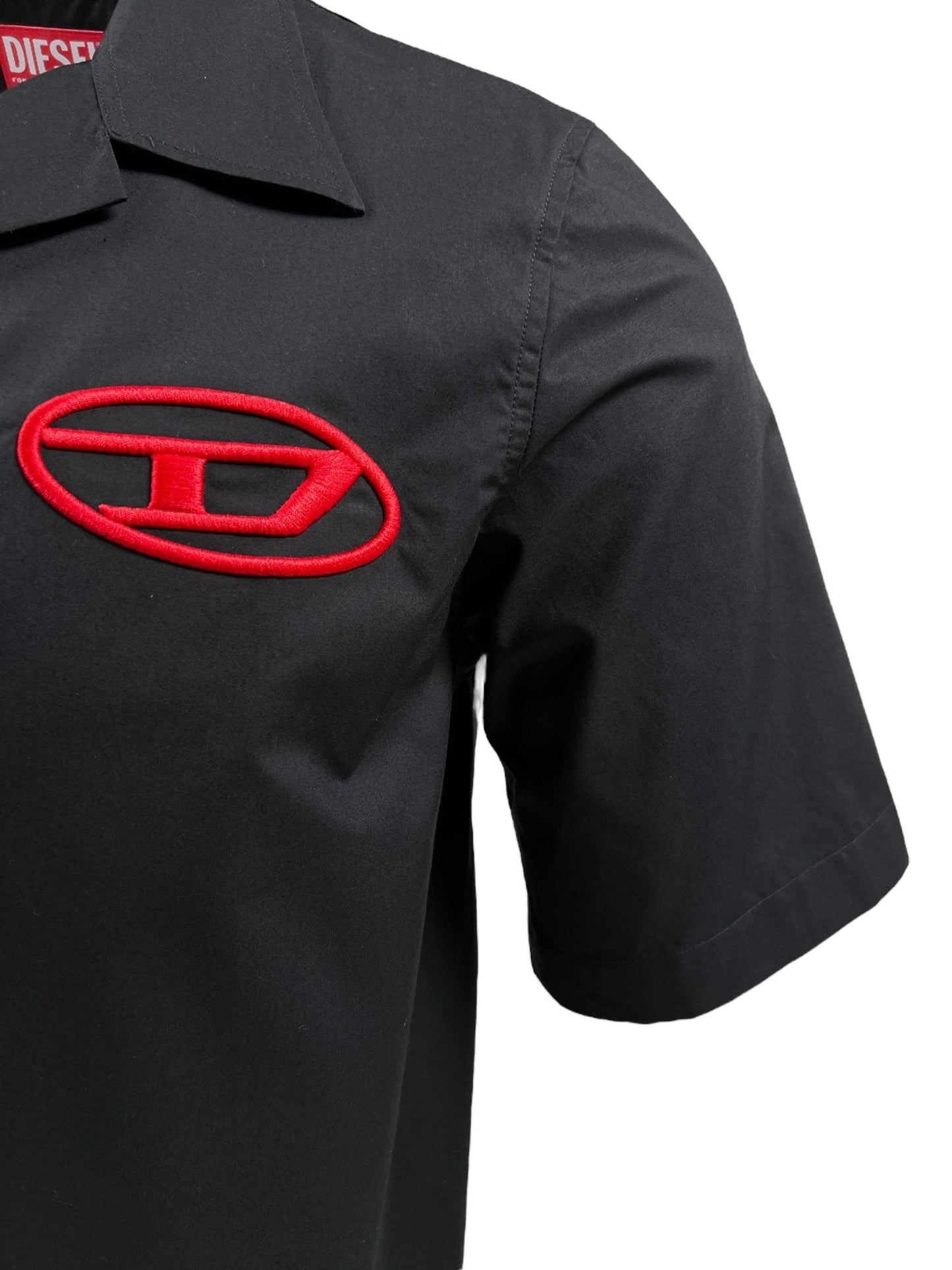 Close-up of the DIESEL S-MAC-C SHIRT BLACK, a short-sleeve men's shirt crafted from organic cotton. It features a relaxed fit and a red embroidered oval logo on the chest, showcasing a stylized "D" inside the oval.