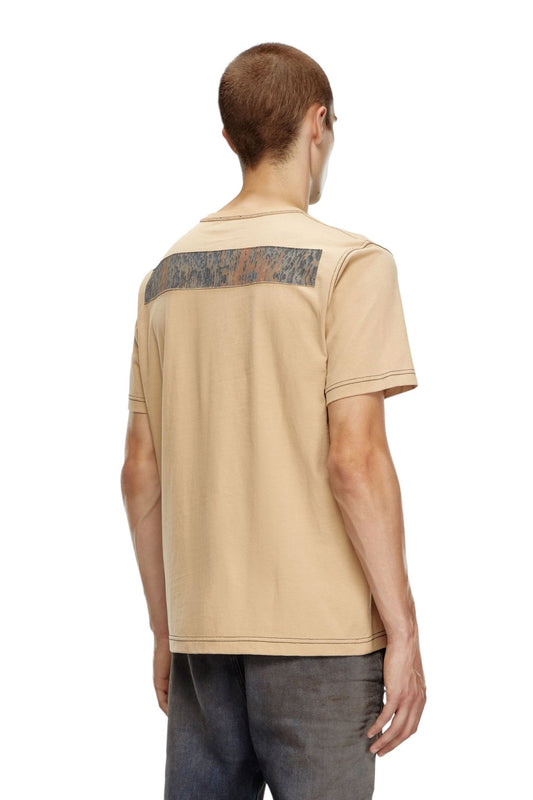 A person with short hair is standing with their back to the camera, wearing the DIESEL T-ADJUST-Q1 T-SHIRT 72S, a beige regular-fit men's cotton T-shirt from DIESEL featuring a rectangular design on the back and dark pants.