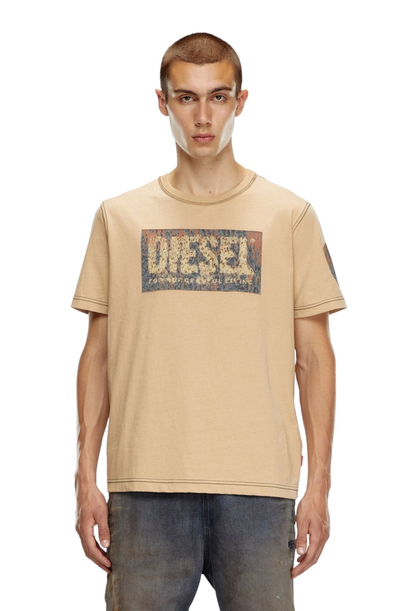 A young man stands facing forward, wearing a regular fit beige DIESEL T-ADJUST-Q1 T-shirt with a distressed "Diesel" logo and faded jeans.