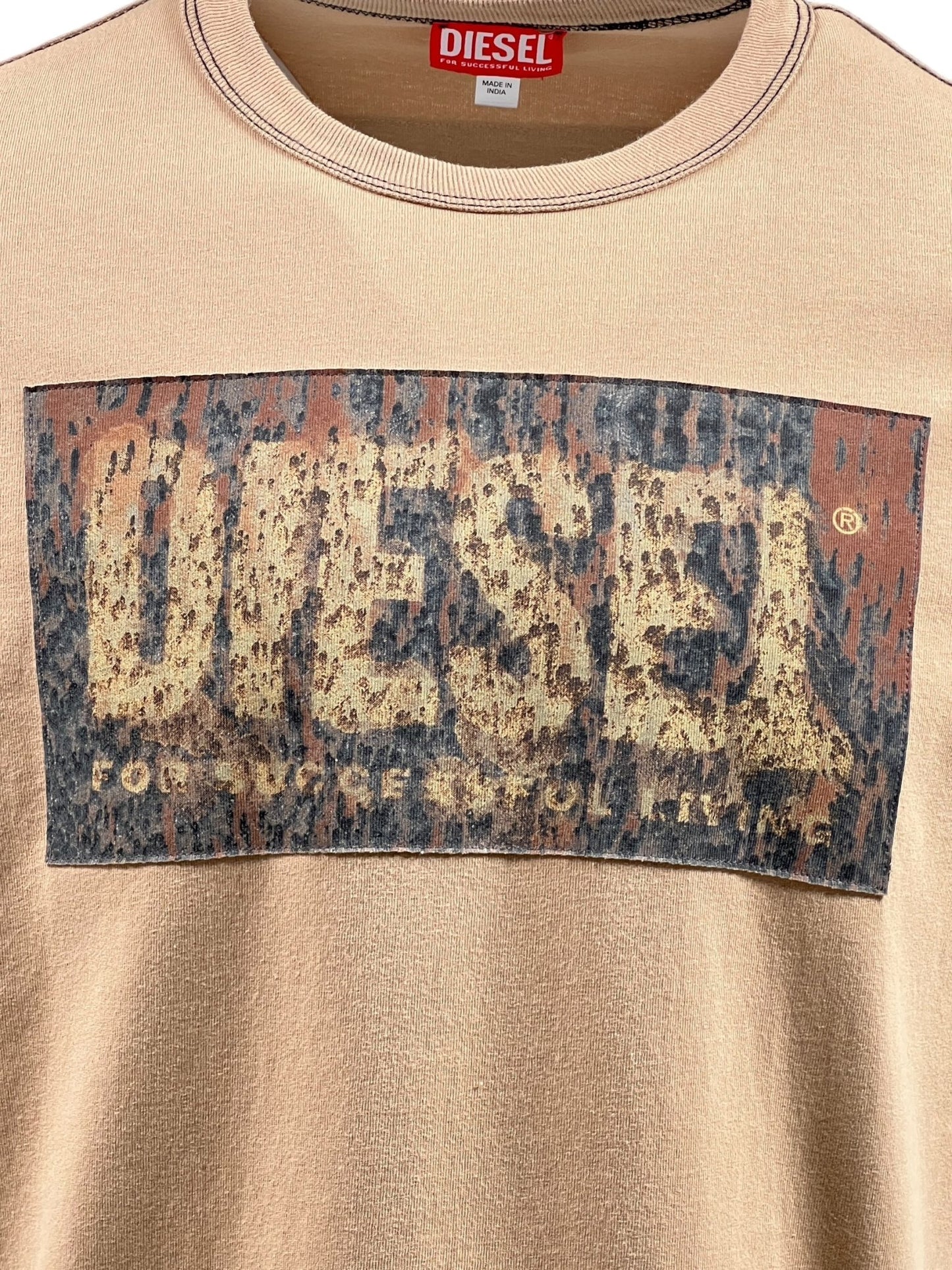 A close-up of the DIESEL T-ADJUST-Q1 T-SHIRT 72S, a beige men’s cotton t-shirt featuring a distressed DIESEL logo on the chest with a unique acid rain effect and a red DIESEL tag on the neckline.