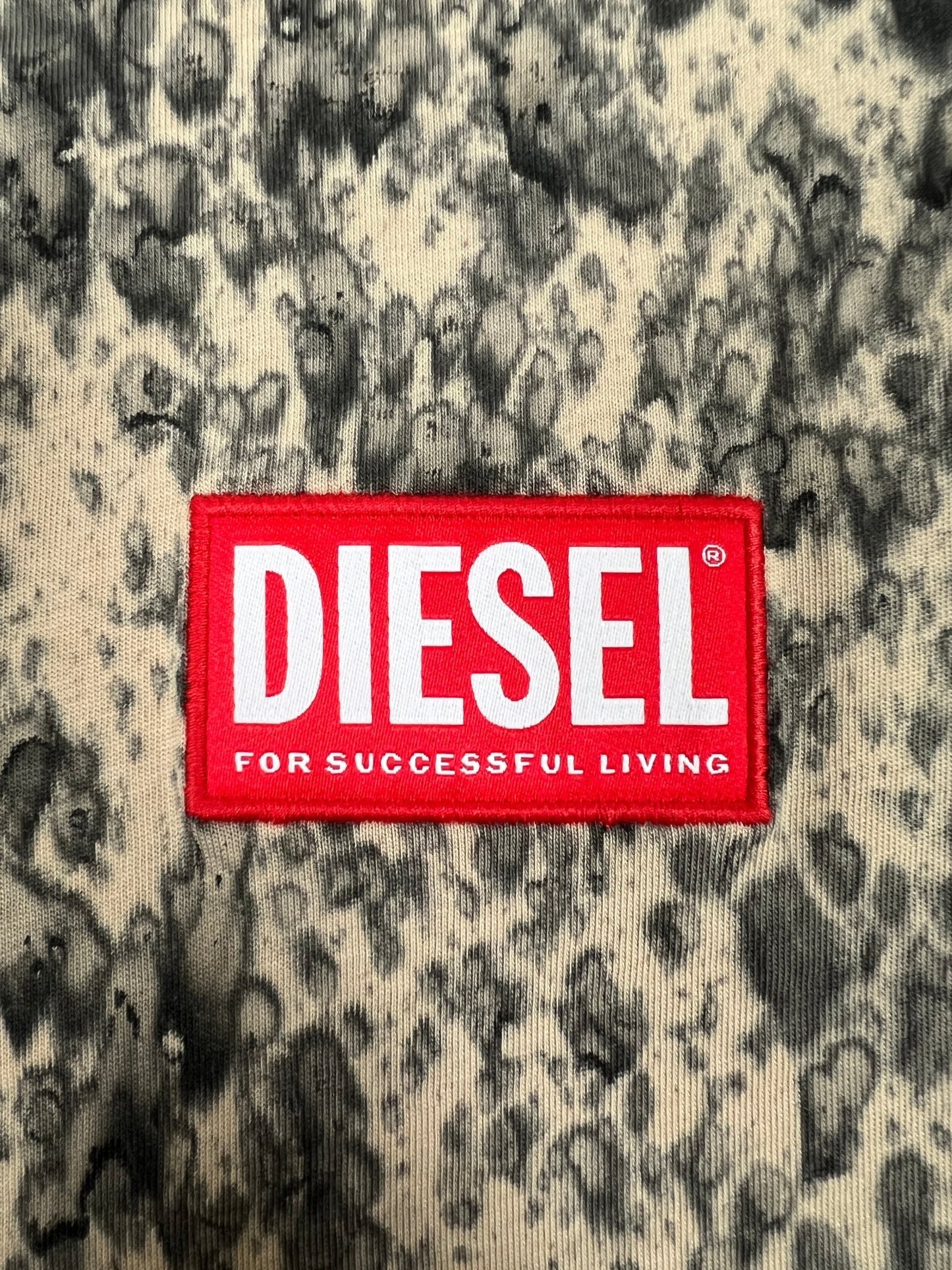 Close-up of a DIESEL T-BOXT-K6 T-SHIRT LIGHT BROWN tag on a textured fabric background. The red tag with white text reads "DIESEL FOR SUCCESSFUL LIVING," reflecting the brand's vibrant and edgy design.