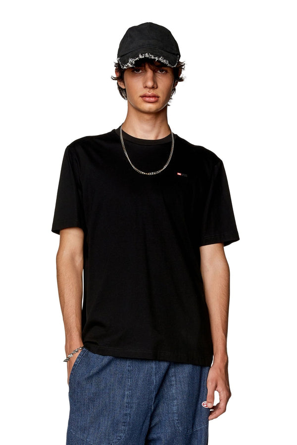 A person wearing a Diesel T-Just-Microdiv T-Shirt in black, paired with a black cap and blue pants from the Diesel Denim Division, stands against a white background.