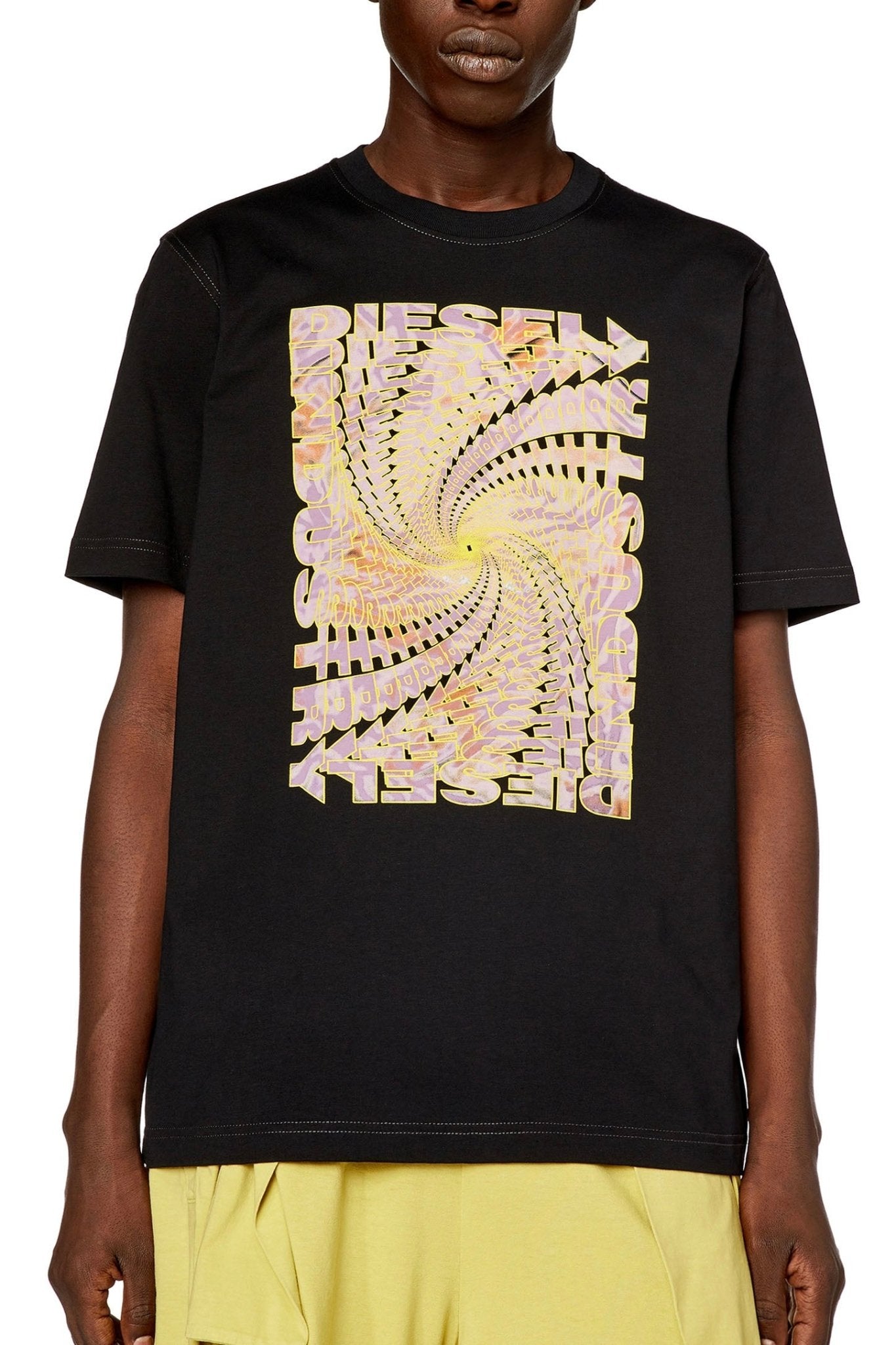 A person wearing a DIESEL T-JUST-N12 T-SHIRT BLACK made from organic cotton jersey, with a graphic spiral design and the word "Diesel" in large, distorted letters from Diesel Industry, paired with yellow shorts.