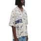 A person wearing a beige Domrebel Bearing camp collar shirt by DOM REBEL and faded jeans stands against a white background.