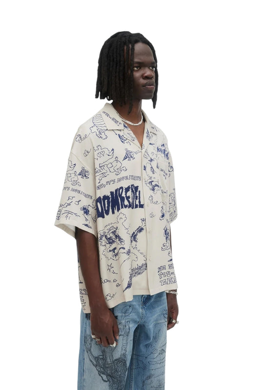 A person wearing a beige Domrebel Bearing camp collar shirt by DOM REBEL and faded jeans stands against a white background.