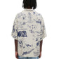 A person with dreadlocks wears a Domrebel Bearing Camp Collar Shirt in beige, featuring playful blue cartoon illustrations and text, paired stylishly with blue jeans. The look is effortlessly set against a plain white background.