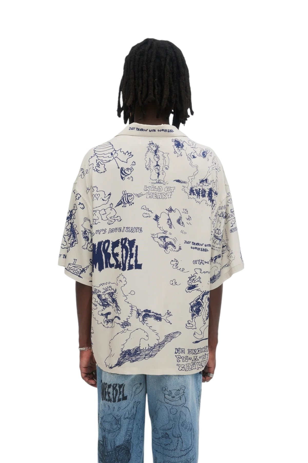 A person with dreadlocks wears a Domrebel Bearing Camp Collar Shirt in beige, featuring playful blue cartoon illustrations and text, paired stylishly with blue jeans. The look is effortlessly set against a plain white background.