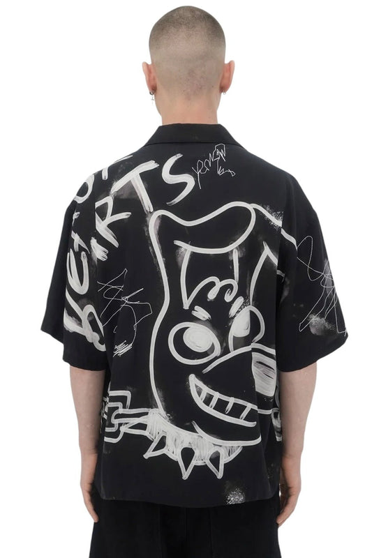 A person with a shaved head is wearing a DOM REBEL Domrebel Chained Shirt in black, which boasts a large abstract, cartoonish graphic design and graffiti elements on the back, made from a soft rayon Tencel blend.