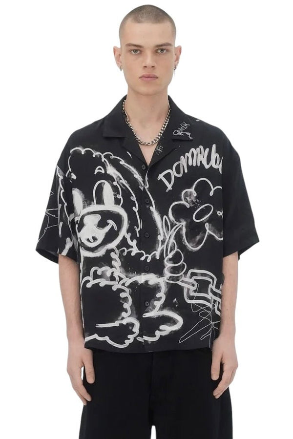 The person is wearing a Domrebel Chained Shirt Black by DOM REBEL, which showcases an all-over print with whimsical white cartoon graphics of a dog and flower on a black background, set against a simple white backdrop.