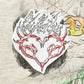 The Domrebel Conflict T-shirt Greige from Dom Rebel features the iconic doodle graphic, displaying a sketched red heart with flame-like edges and horns amidst abstract scribbles and shapes.