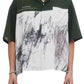 The person is wearing the Domrebel Demi Camp Collar Shirt by DOM REBEL, featuring an abstract pattern in dark green and white with "PAINTINGS and DRAWINGS" text. Made from twill fabric, it has a loose fit, complemented by various jewelry pieces.