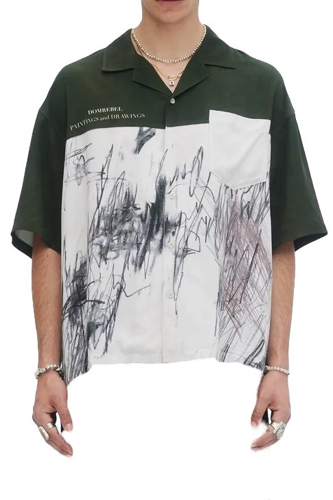 The person is wearing the Domrebel Demi Camp Collar Shirt by DOM REBEL, featuring an abstract pattern in dark green and white with "PAINTINGS and DRAWINGS" text. Made from twill fabric, it has a loose fit, complemented by various jewelry pieces.