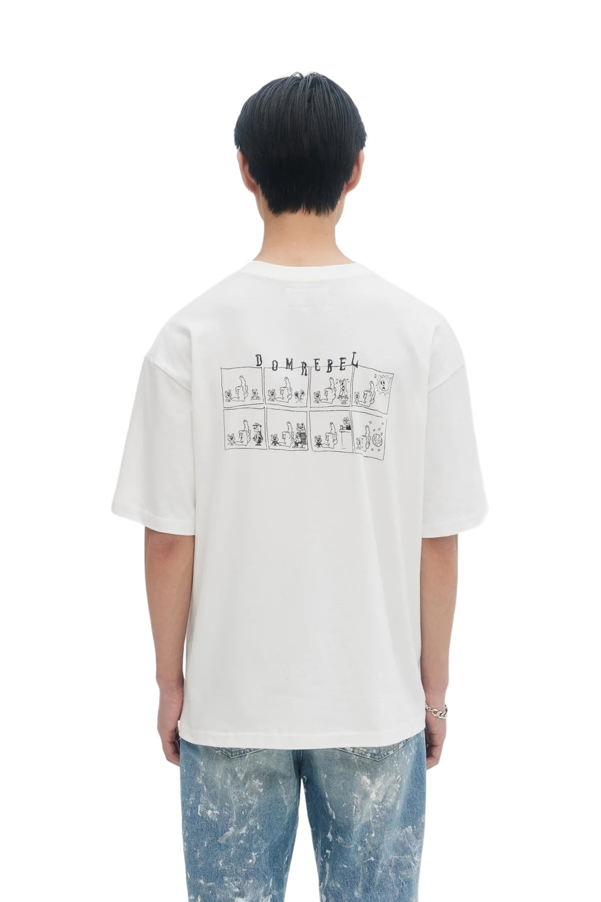 A person wearing a Domrebel Friendly T-Shirt Ivory with a comic-style teddy bear pairs it with blue splattered jeans, viewed from the back—perfect for those who love the eclectic style of the DOM REBEL bon vivant collection.