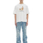 A person stands in an ivory Domrebel Friendly T-Shirt by DOM REBEL, featuring hand-drawn teddy bears. They're paired with blue splattered jeans and sneakers against a plain white background.