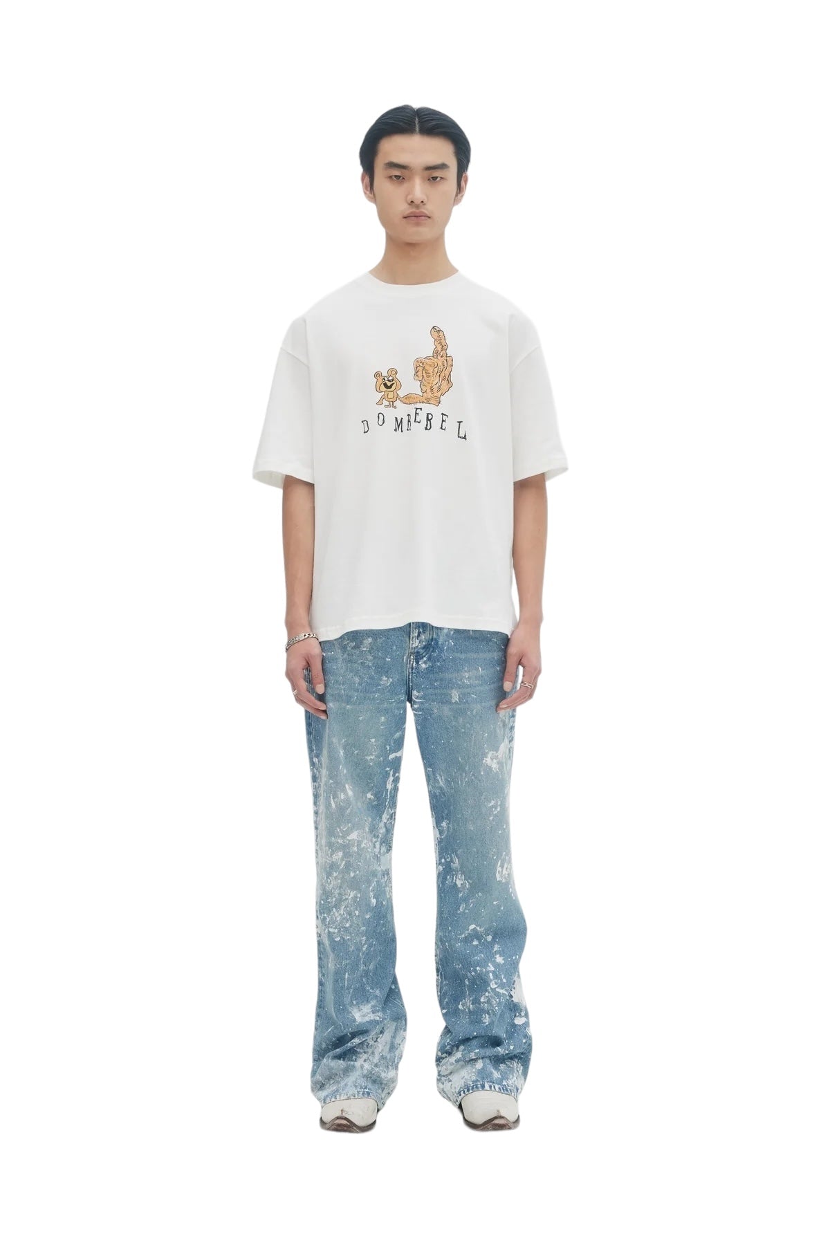A person stands in an ivory Domrebel Friendly T-Shirt by DOM REBEL, featuring hand-drawn teddy bears. They're paired with blue splattered jeans and sneakers against a plain white background.