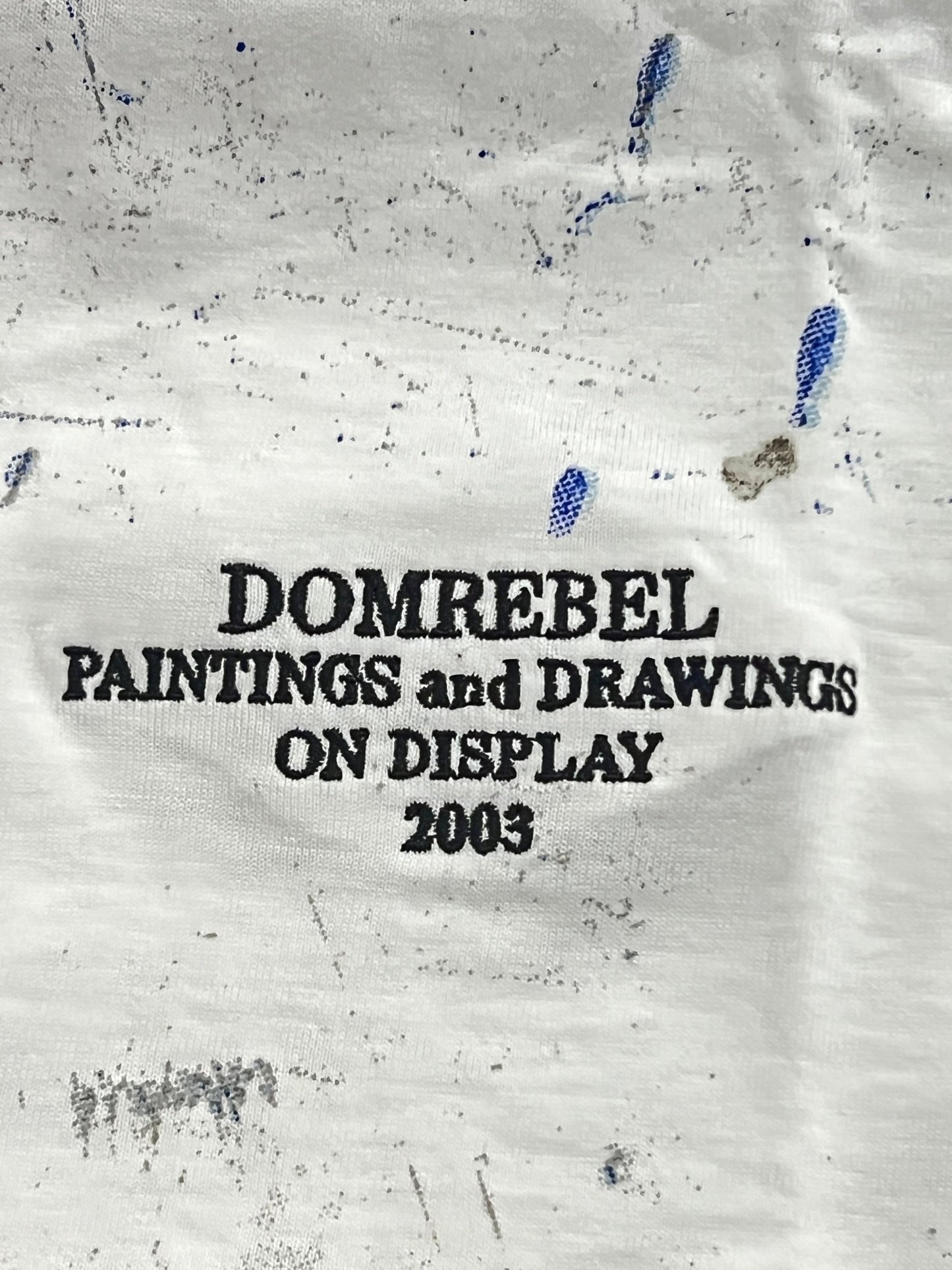 Close-up of a DOM REBEL DOMREBEL RAG T-SHIRT IVORY made from 200GSM RINGSPUN JERSEY COTTON with paint splatters, featuring black embroidered text that reads "DOMREBEL PAINTINGS and DRAWINGS ON DISPLAY 2003.