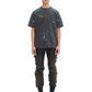 Against a white background, a person wears the DOM REBEL Domrebel Atelier Pocket Tee Coal Paint, paired with cargo pants and black boots.