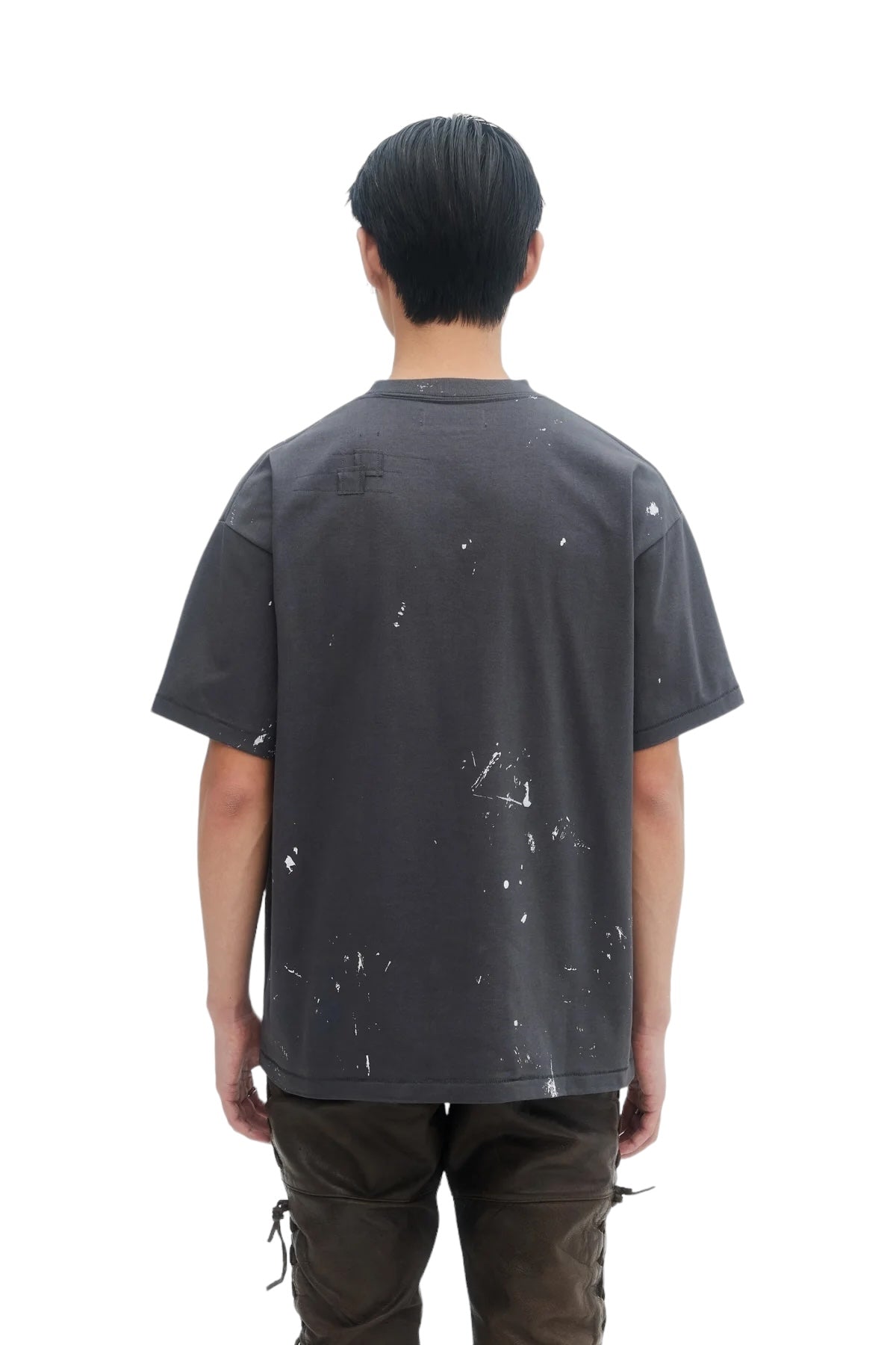 A person with dark hair stands facing away, wearing a DOM REBEL Domrebel Atelier Pocket Tee Coal Paint with a unique white splatter design and dark trousers.