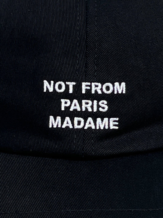 Close-up of a black cotton baseball cap by DROLE DE MONSIEUR, featuring the embroidered text "NOT FROM PARIS MADAME" in white. The product name is CP151-CO138-BL LA CASQUETTE SLOGAN BLK.