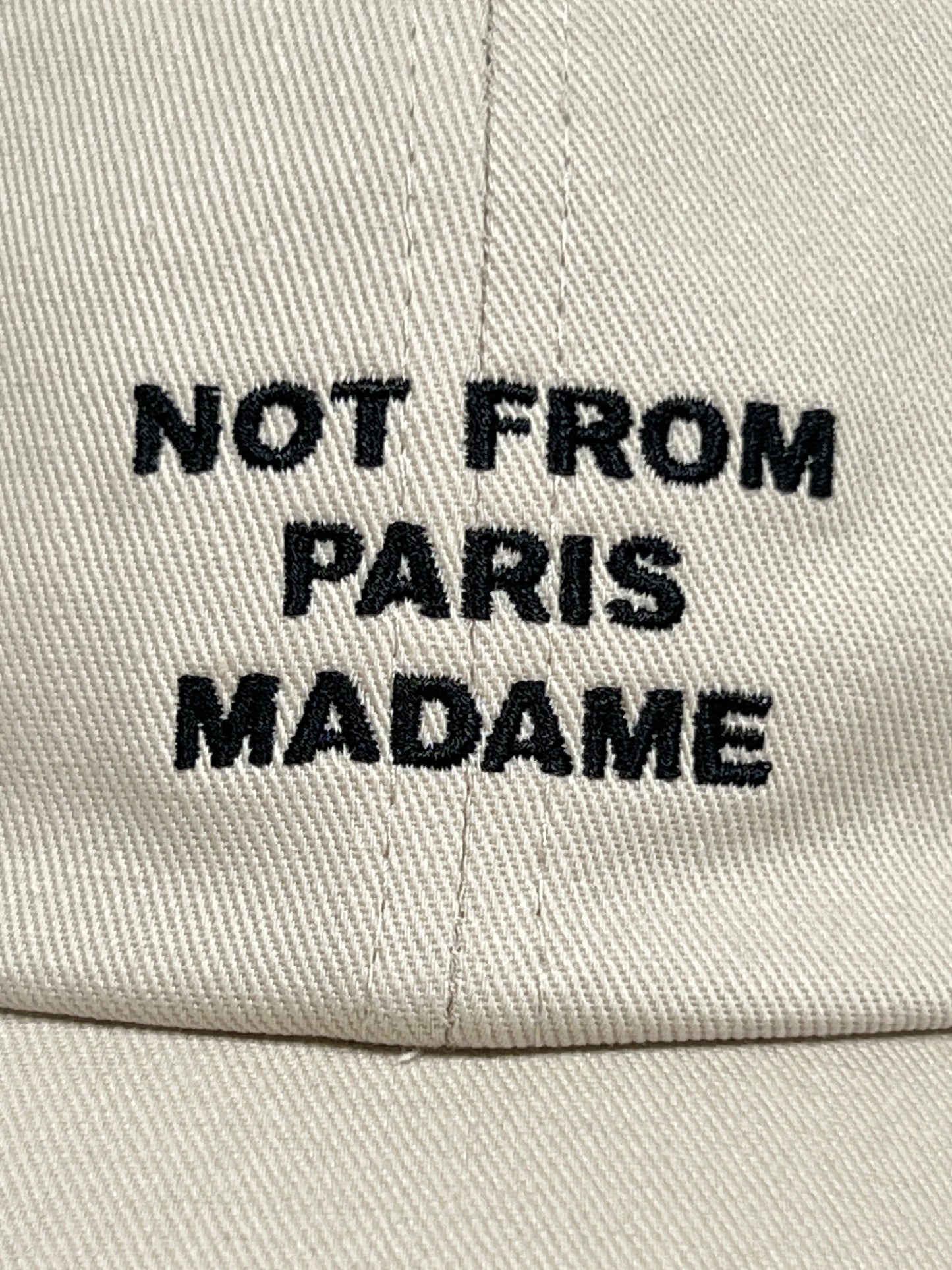 Beige fabric baseball hat with an adjustable strap, featuring black embroidered text reading "NOT FROM PARIS MADAME" DROLE DE MONSIEUR CP151-CO138-MT LA CASQUETTE SLOGAN MASTIC from DROLE DE MONSIEUR.