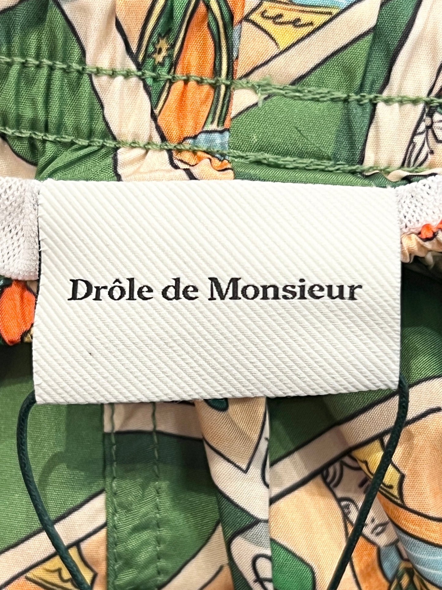 Clothing label with the text "DROLE DE MONSIEUR" attached to mid-length swim shorts featuring a card game print. (Product Name: DROLE DE MONSIEUR D-BS141-PL136 LE SHORT PLAGE JEU DE CARTES GN)