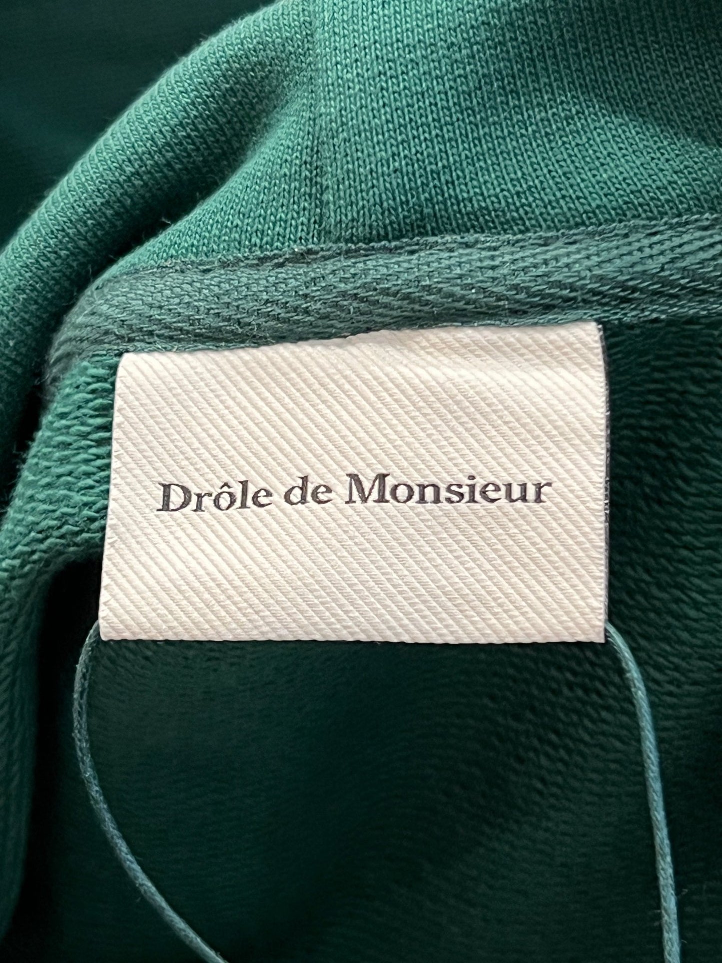 Close-up of a DROLE DE MONSIEUR D-HO146-CO127-FGN LE HOODIE NATURE MORTE clothing label on a French terry sweatshirt fabric background.